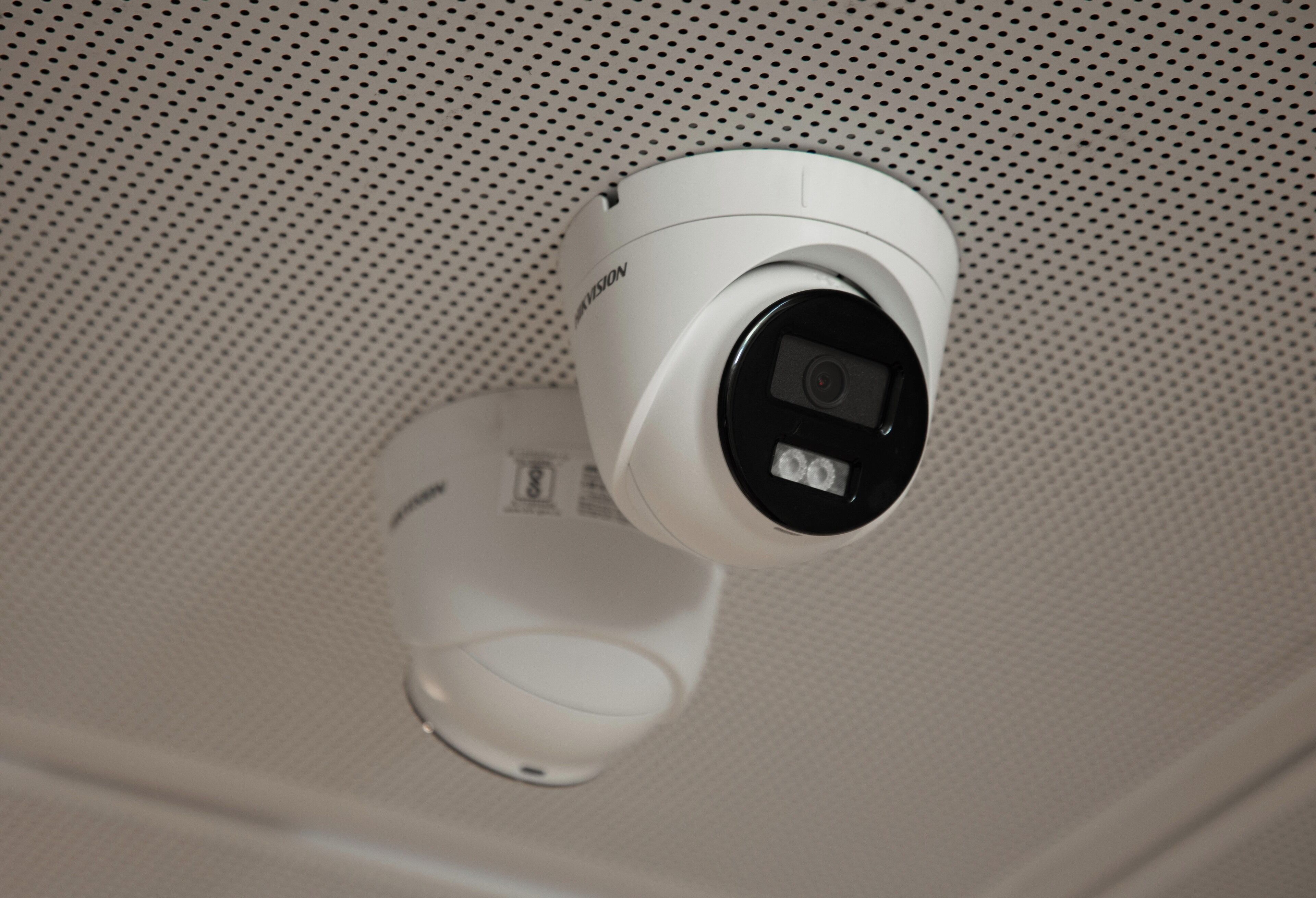A white security camera mounted on a ceiling with a perforated pattern.