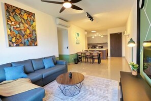 A modern living room with a sofa, coffee table, and a ceiling fan. A kitchen is visible in the background with a dining area. - M Vertica KL City 4 Bedroom Fully Air-Con Suite (Kuala Lumpur)