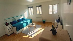 A bedroom with a bed, bedside table, and a small desk with a chair. There are windows and a radiator on the wall. - New Studio/Near Airport CDG/Direct Paris center/Disneyland/Villepinte EXPO (Tremblay En France)