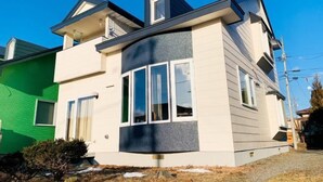 A two-story house with a dark roof, light siding, and a prominent corner window. - Pokke (Shiraoi)