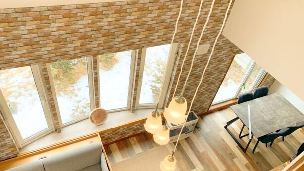 A high-ceiling room with a brick wall, large windows, a sofa, a dining table, and hanging pendant lights. - Pokke (Shiraoi)