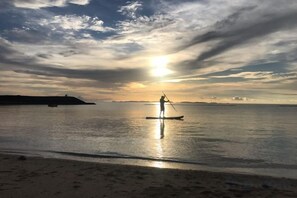A beach scene with a person paddleboarding near the shore, calm water, and a sunset in the background. - ZZOKIAI Blue Fish! Only 2 mins to beach by walk! Maximun 11 persons! (Itoman)