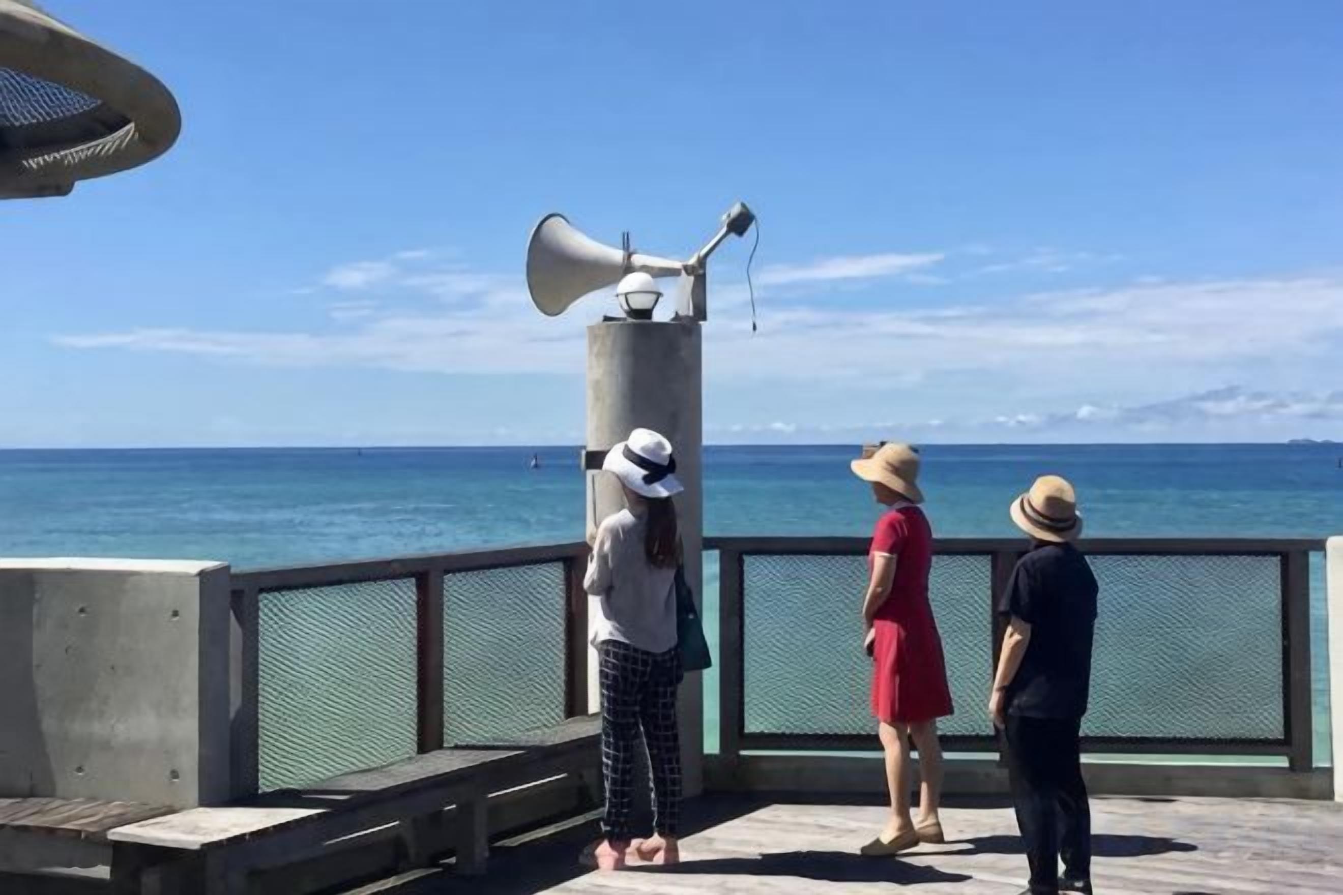 Um píer com grade de metal, um pilar de concreto com um megafone e uma vista ampla do oceano.