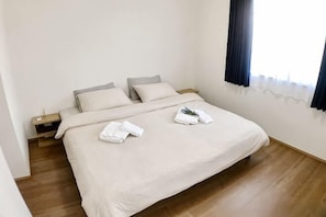 A neatly made bed with a beige bedspread and pillows, a wooden floor, and a nightstand with a lamp.