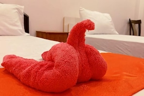 Un peluche rojo sobre una manta naranja en una habitación de hotel con ropa de cama blanca y una mesita de noche de madera.