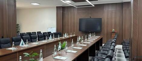 A conference room with a long table, multiple chairs, a flat-screen TV, and a plant.