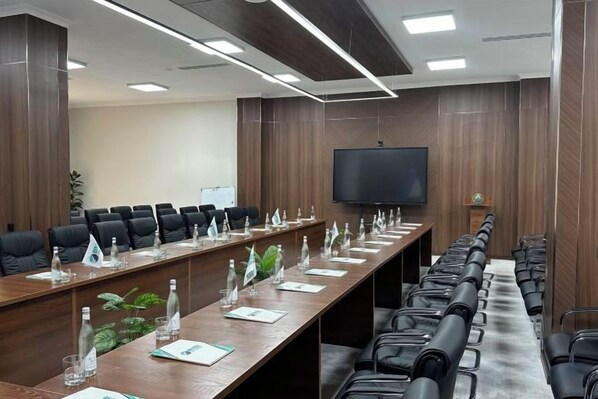 A conference room with a long table, multiple chairs, a flat-screen TV, and a plant.
