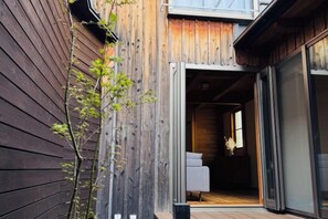 Fachada de madera con puerta corrediza de vidrio que da acceso a un espacio interior con sofá y ventana.