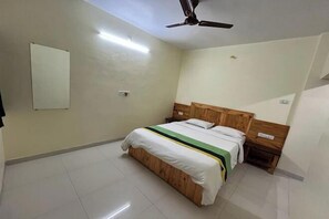 A bedroom with a wooden bed, white bedding, a ceiling fan, and a small bedside table. - Savitri Bungalow (Dhangarwadi)