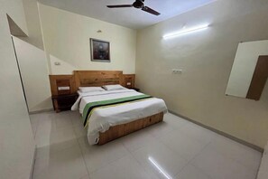 A bedroom with a wooden bed, a ceiling fan, a nightstand, a wall-mounted mirror, and a painting. - Savitri Bungalow (Dhangarwadi)