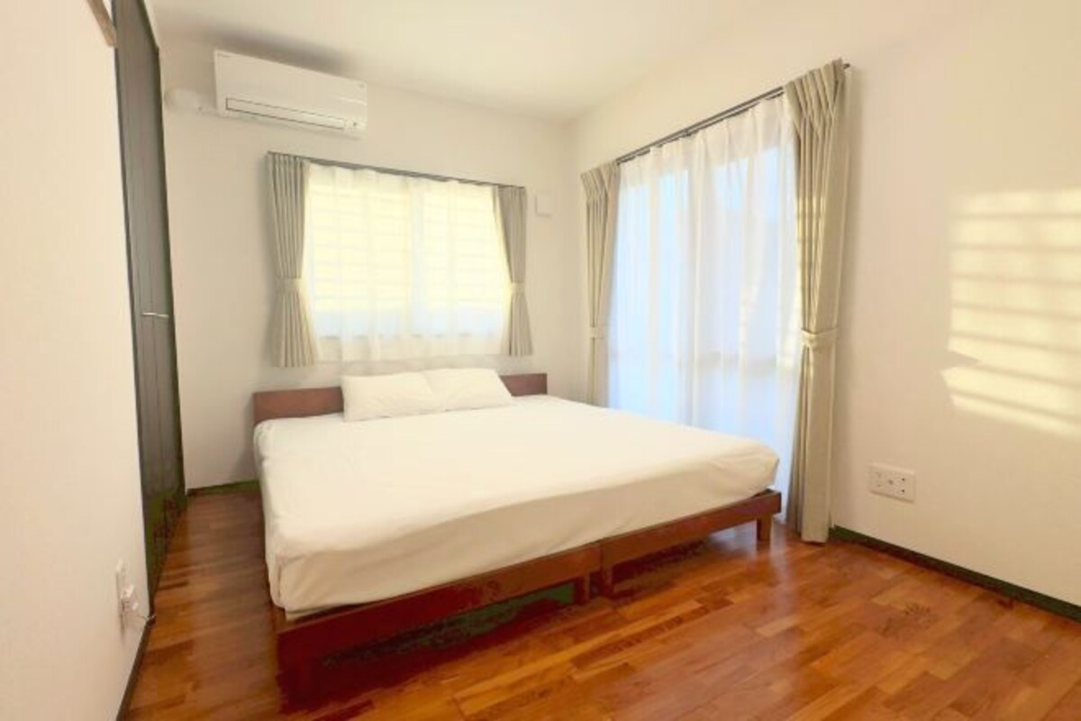 A single bed in a room with wooden flooring, a window with curtains, and an air conditioning unit on the wall.