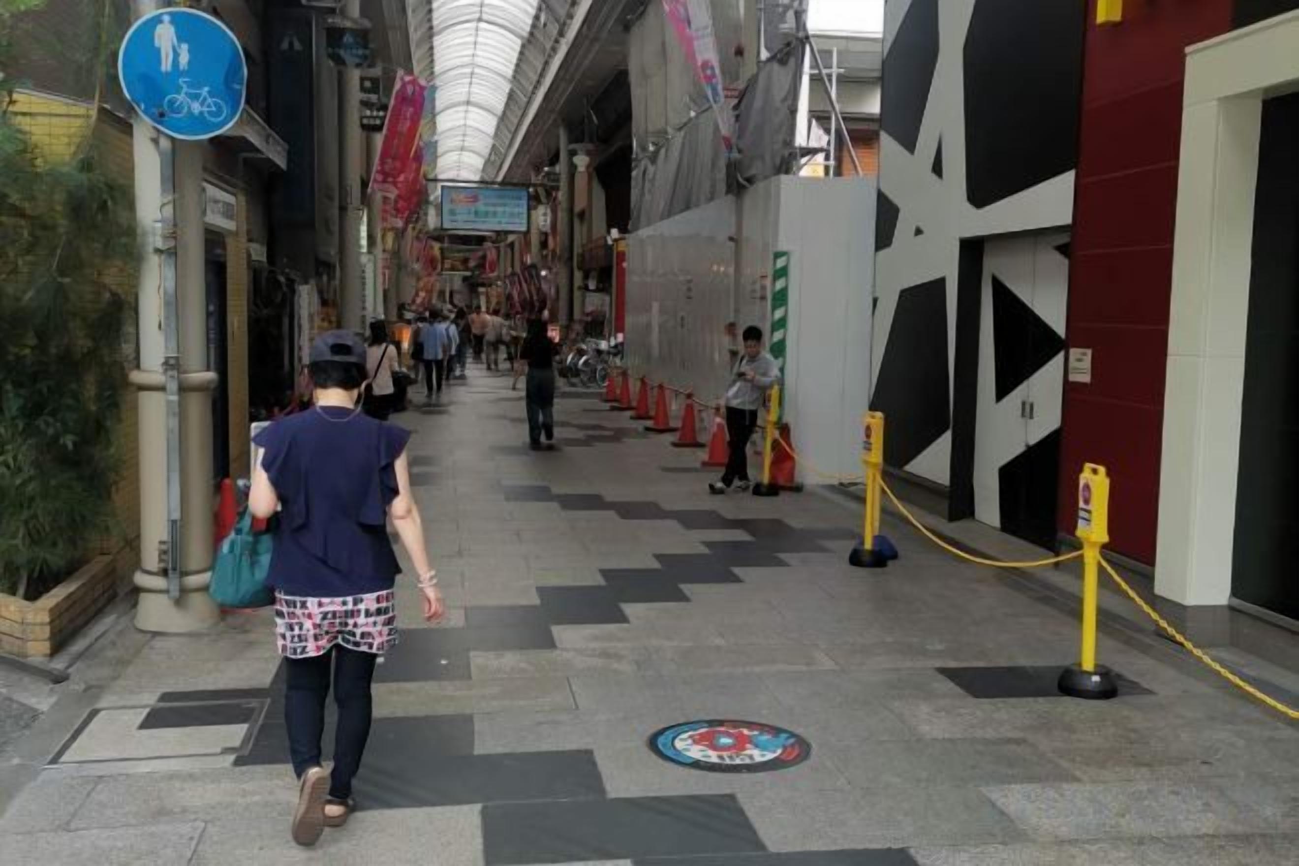 A pedestrian walkway with a blue sign indicating a bicycle route, a red and white building, and yellow stanchions.
