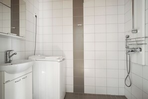 A modern bathroom with white tiles, a sink, a toilet, and a shower area. - Kotimaailma Aparthotel Kamppi - 1BR for 3 (Helsinki)