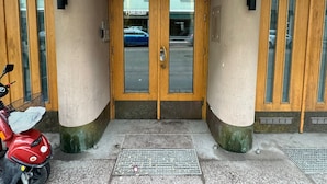 Entrada de um edifício com uma placa indicando o número 31 acima das portas, uma scooter vermelha estacionada na frente e uma grade de metal na entrada.