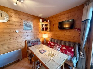 Interior acolhedor de cabana com parede de madeira, mesa com colcha, sofá com manta de pele, TV fixada na parede e armário com objetos decorativos.