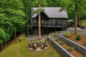 A cabin with a deck, surrounded by trees, and a fire pit with chairs.