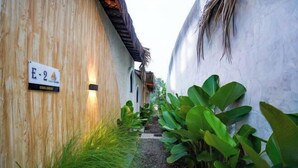 Un étroit sentier bordé de murs en bois et d’une végétation luxuriante, qui mène à un bâtiment sur lequel est apposée une plaque indiquant « E-2 ».