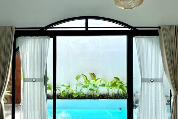 Habitació amb finestra gran que dona a una piscina i vegetació.