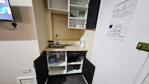 Eine kleine Küchenzeile mit Spüle, Wasserkocher und einem Hängeschrank. Eine Mülltonne und eine Steckdose befinden sich an der Wand.