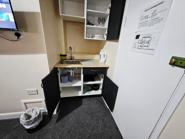 A small kitchenette with a sink, a kettle, and a cabinet. There is a trash bin and a power outlet on the wall.