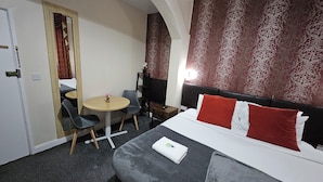Ein Hotelzimmer mit Bett, Schreibtisch und Stuhl. Ein kleiner Tisch mit Blumenvase steht darin. Die Wände sind mit einem gemusterten Wandteppich versehen, und ein Vorhang drapiert sich vor der Tür.