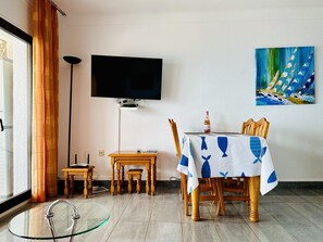 A dining area with a table set for two, a television mounted on the wall, and a painting above the table.