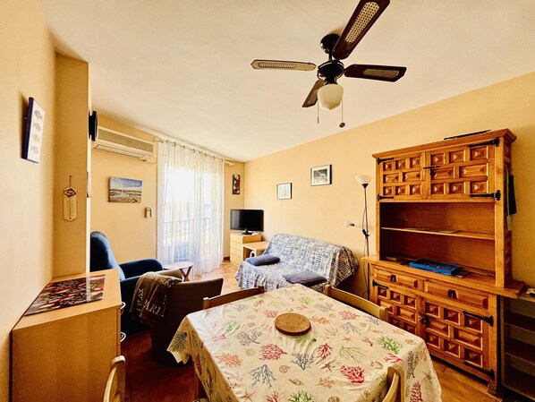 A room with a ceiling fan, a television, a wooden cabinet, a table with a floral tablecloth, and a chair.