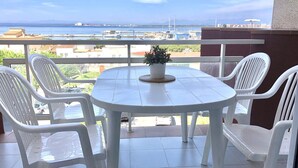 A balcony with a white table and chairs, a potted plant, and a view of the sea and buildings.