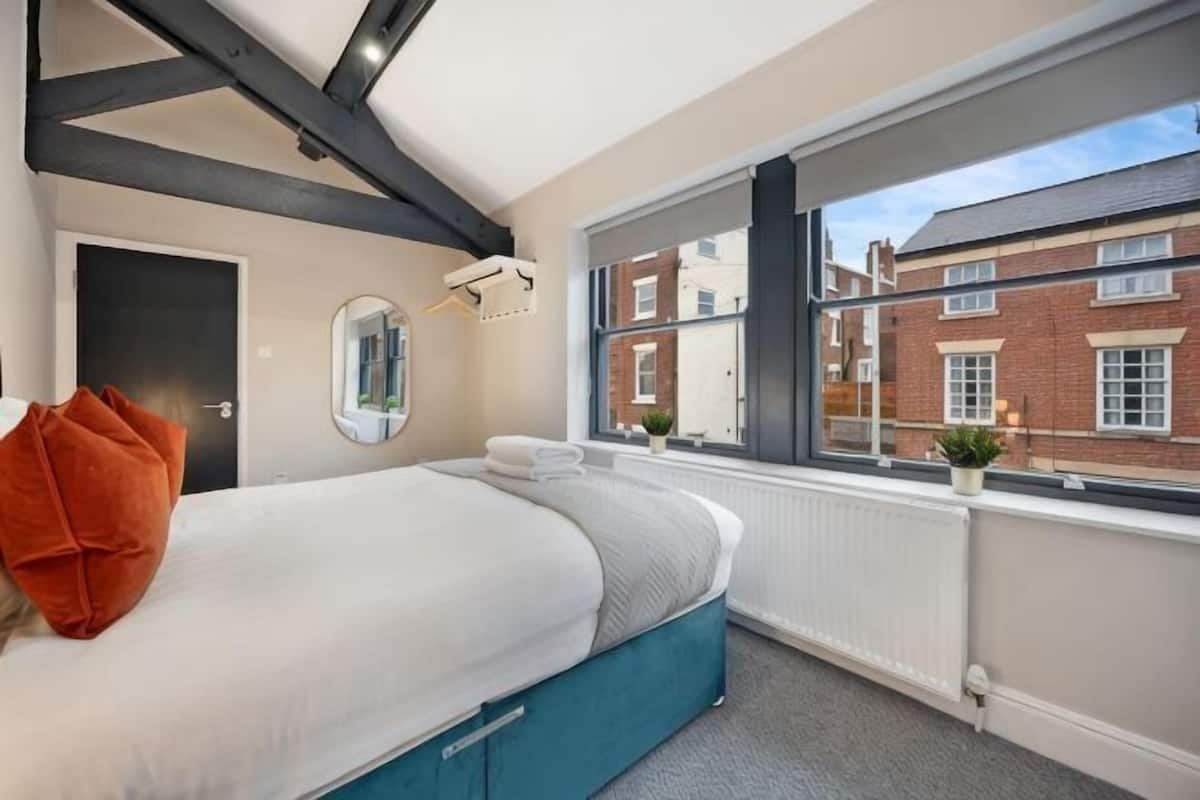 A bedroom with a bed, a window with a view of buildings, a radiator, and a door.