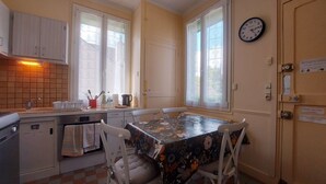 A compact kitchen with a dining table, chairs, a clock, and a window with curtains. - Cozy lodging near Chartres - 6 guests (Mainvilliers)