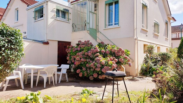 A two-story house with a covered patio, a small outdoor seating area with chairs, and a large flowering bush. - Cozy lodging near Chartres - 6 guests (Mainvilliers)