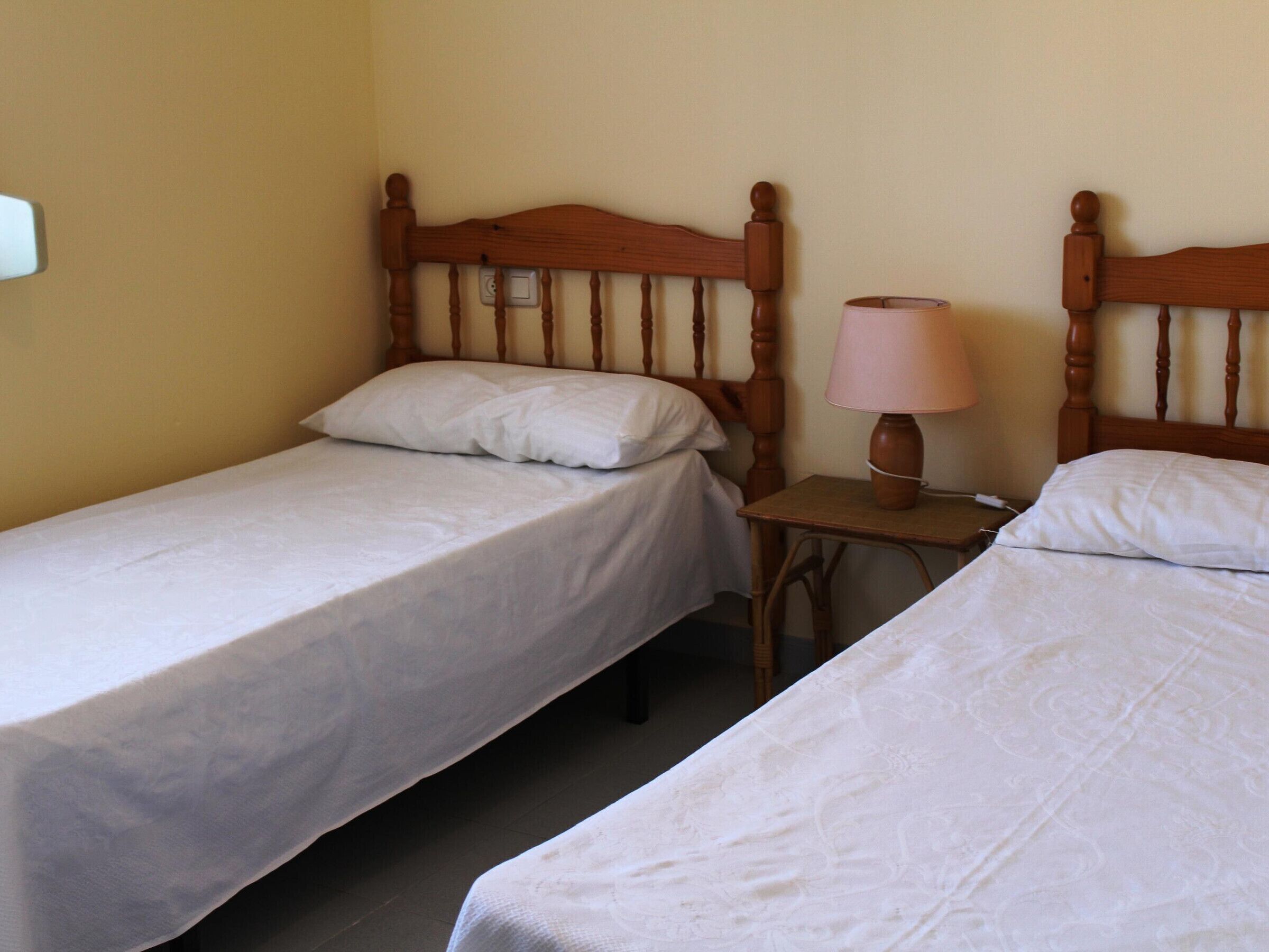A room with two single beds, wooden headboards, a bedside table with a lamp, and a wall socket.