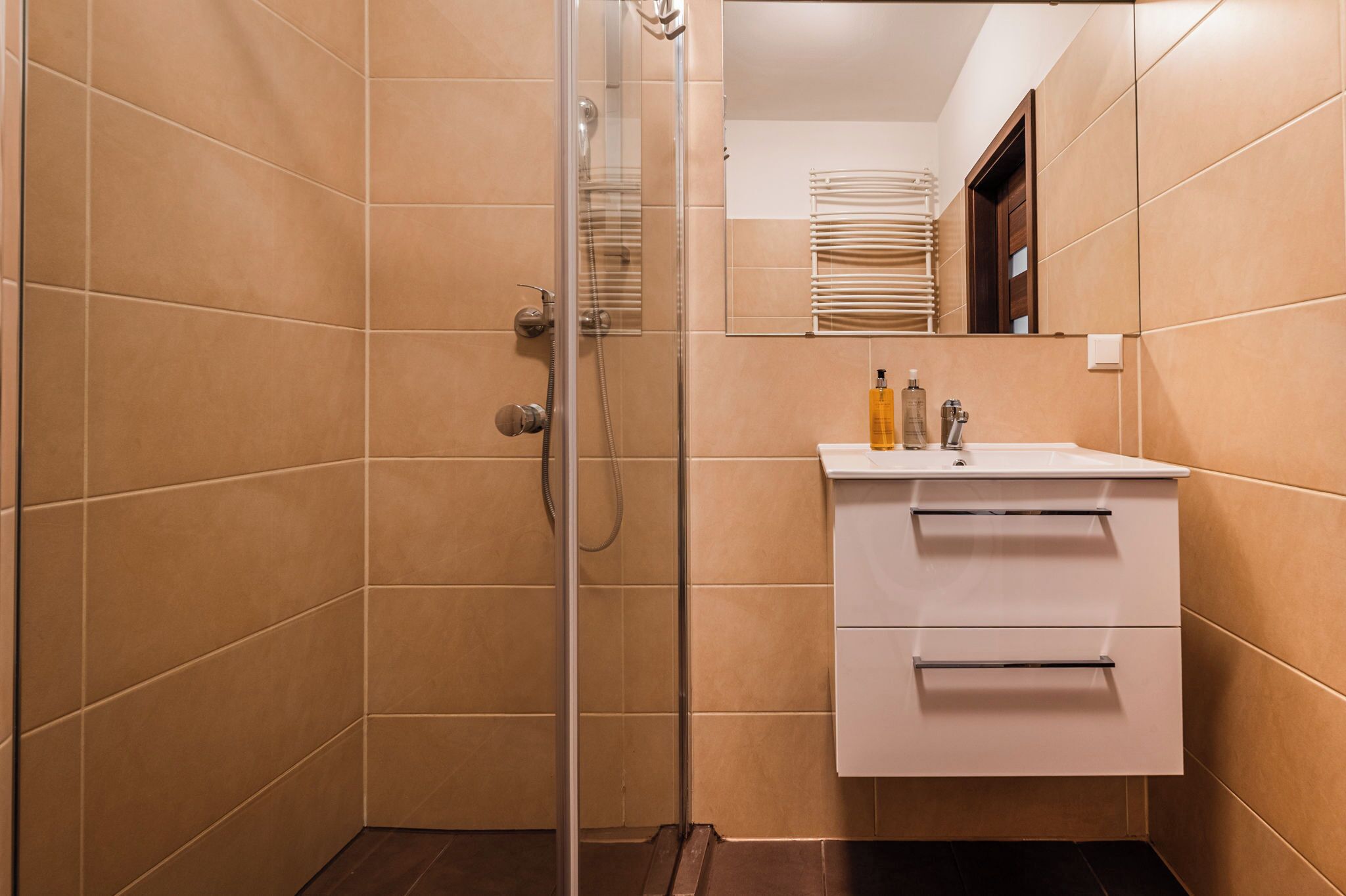 Un baño con paredes de azulejos color beige, un box de ducha de vidrio, un lavamanos blanco con un espejo encima y un perchero visible en el reflejo.