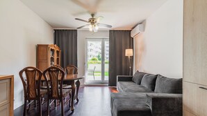 Uma sala de estar moderna com um sofá cinza escuro, mesa de jantar de madeira com cadeiras, ventilador de teto e vista para o exterior através de uma porta de vidro.
