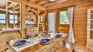 L’intérieur d’un chalet en bois, avec une table à manger dressée pour quatre personnes, des poutres en bois au plafond et une vue sur la verdure à l’extérieur.