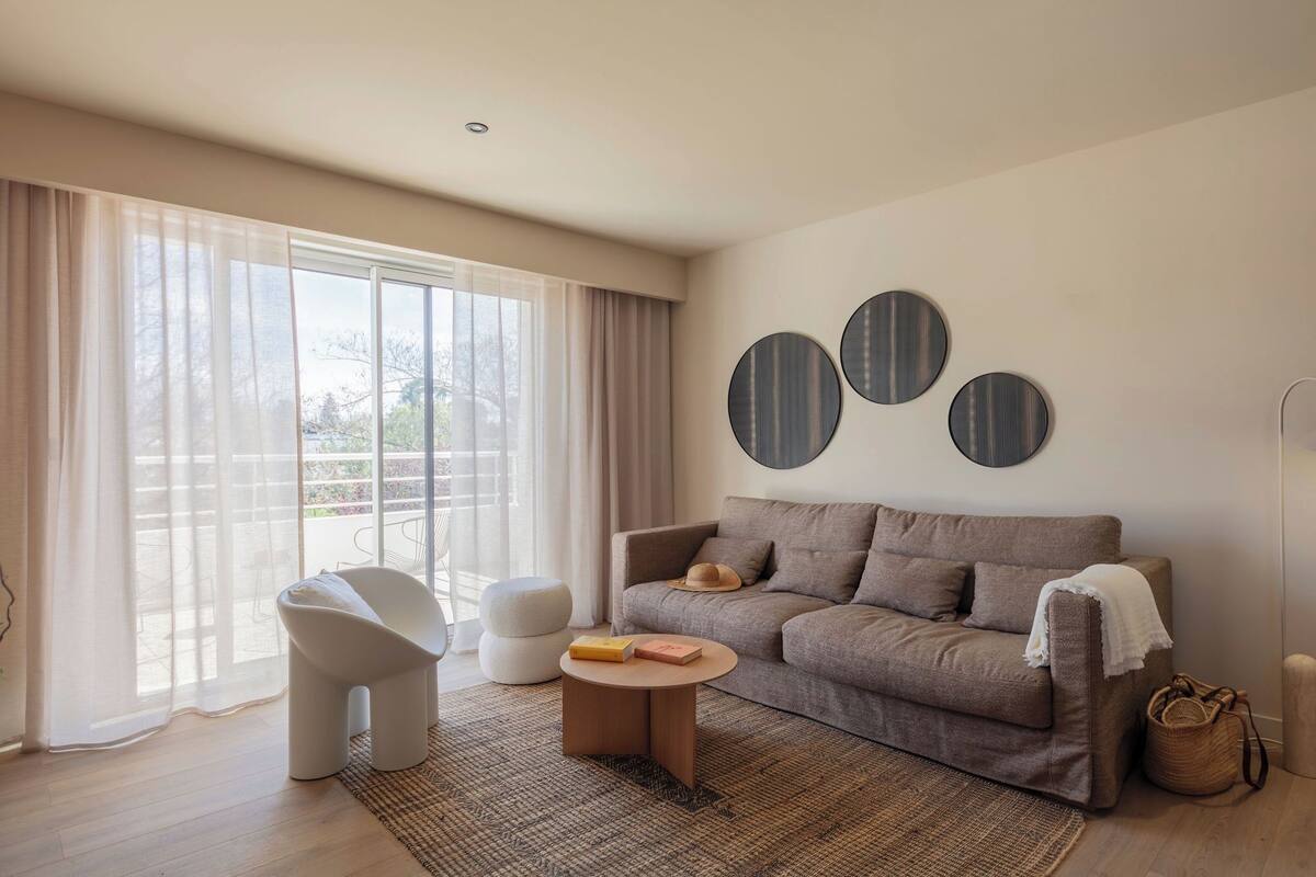 Un salon moderne avec un canapé, un fauteuil, une table basse ronde et de grandes fenêtres ornées de rideaux légers.
