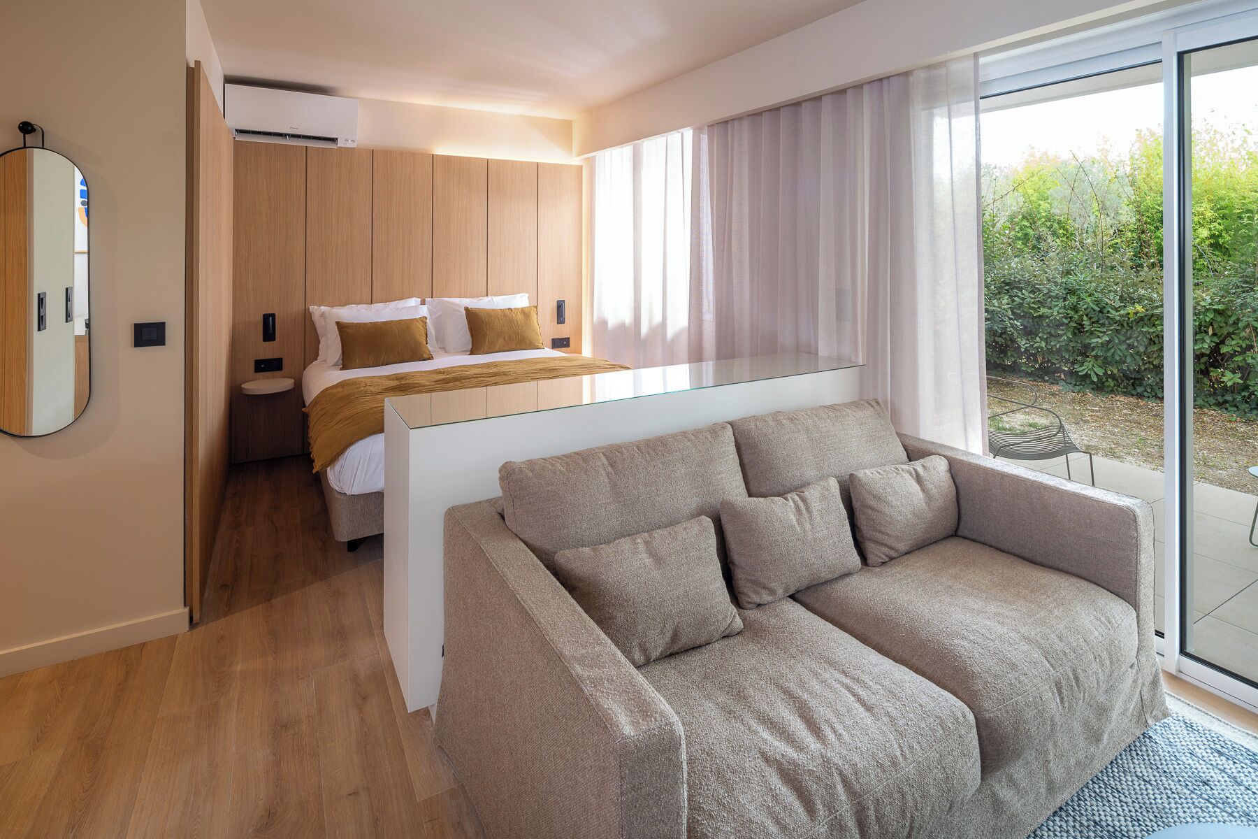Une chambre d’hôtel moderne avec un lit, un canapé, une cloison en verre et une vue sur la verdure.