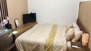 A hotel room with a bed, a TV, a bedside table with a lamp, and a minibar.