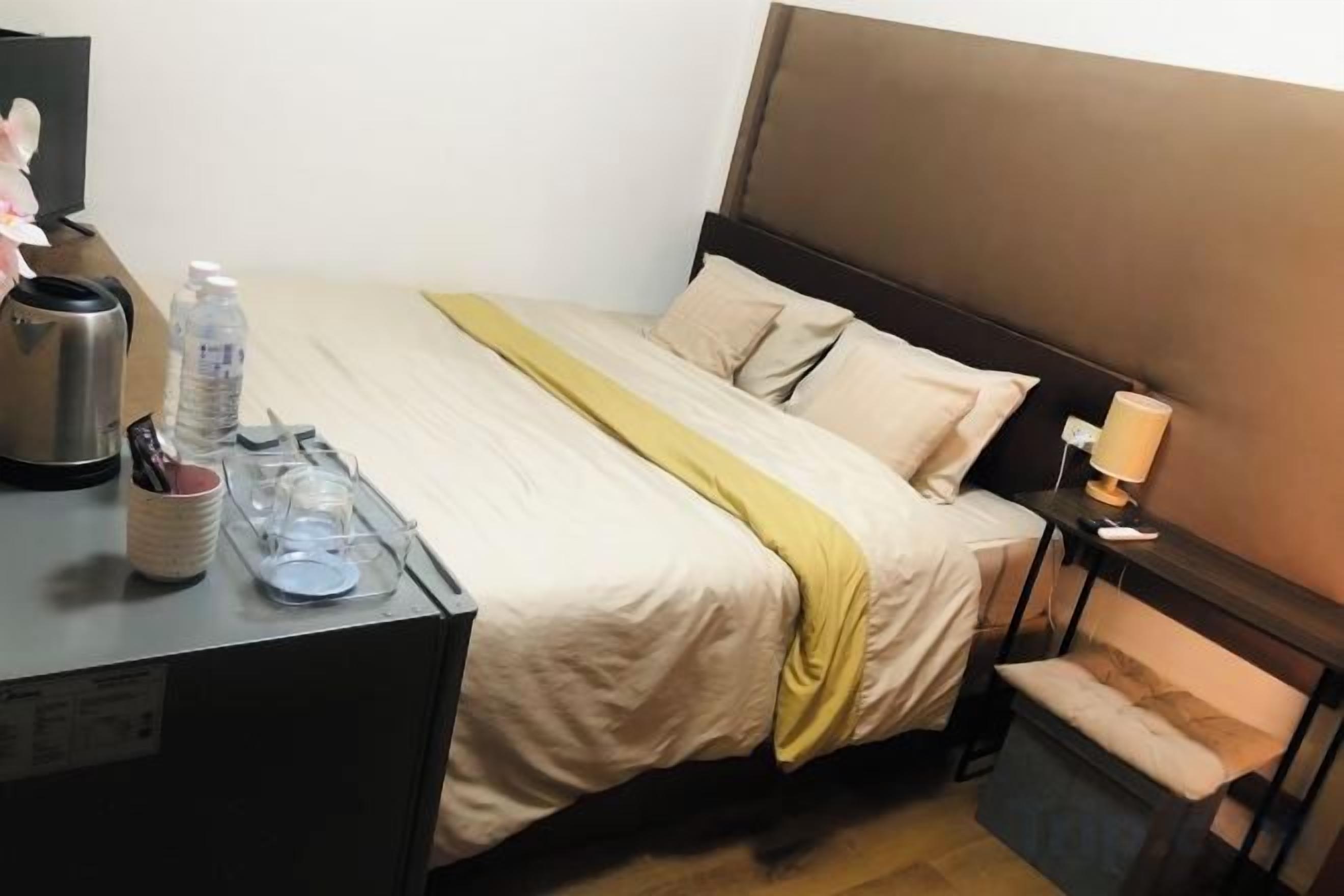 Hotellihuone, jossa on sänky, yöpöytä, lamppu ja pieni työpöytä, sekä vedenkeitin ja vesipullo.
