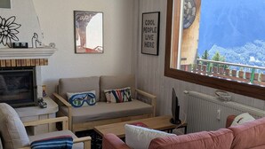 A cozy living room with a fireplace, a sofa, a pink couch, and a view of a snowy mountain through a window.