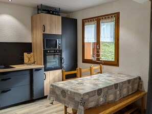 A compact kitchen with a dining area, featuring a table with a patterned tablecloth, a window with curtains, and a view of greenery outside.