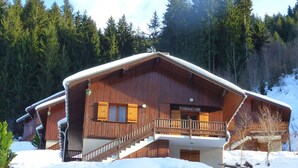 Una cabaña de madera con un balcón y una escalera que conduce a la entrada, rodeada de nieve y árboles.