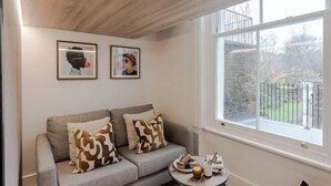 A modern living room with a grey sofa, two patterned cushions, a small round table with a candle, and framed artwork on the wall. - Kensington The Loft XL (London)