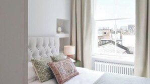 A bedroom with a tufted headboard, a bedside table with a lamp, and a view of buildings through a large window. - B Comfort Apartments (London)