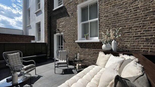 A brick exterior wall, a white cushioned sofa, and two wicker chairs on a patio. - B Comfort Apartments (London)