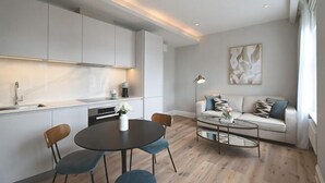 A modern kitchen with white cabinets, a dining area with a round table and chairs, and a living area with a sofa and coffee table. - B Comfort Apartments (London)