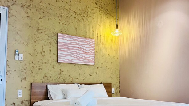Una cama bien hecha con sábanas blancas y una toalla enrollada, un cabecero de madera, una lámpara de pared y una pared con textura.