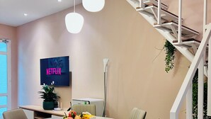 Un comedor moderno con una mesa redonda, sillas, un televisor mostrando 'NETFLIX' y una escalera con una planta.