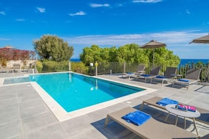 Zona de piscina con sillones de descanso y sombrillas, rodeada de vegetación y vistas al mar.