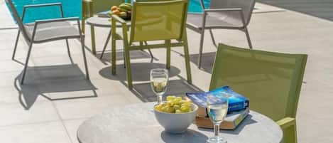 A poolside area with a table, chairs, a bowl of olives, a glass of wine, and a book.